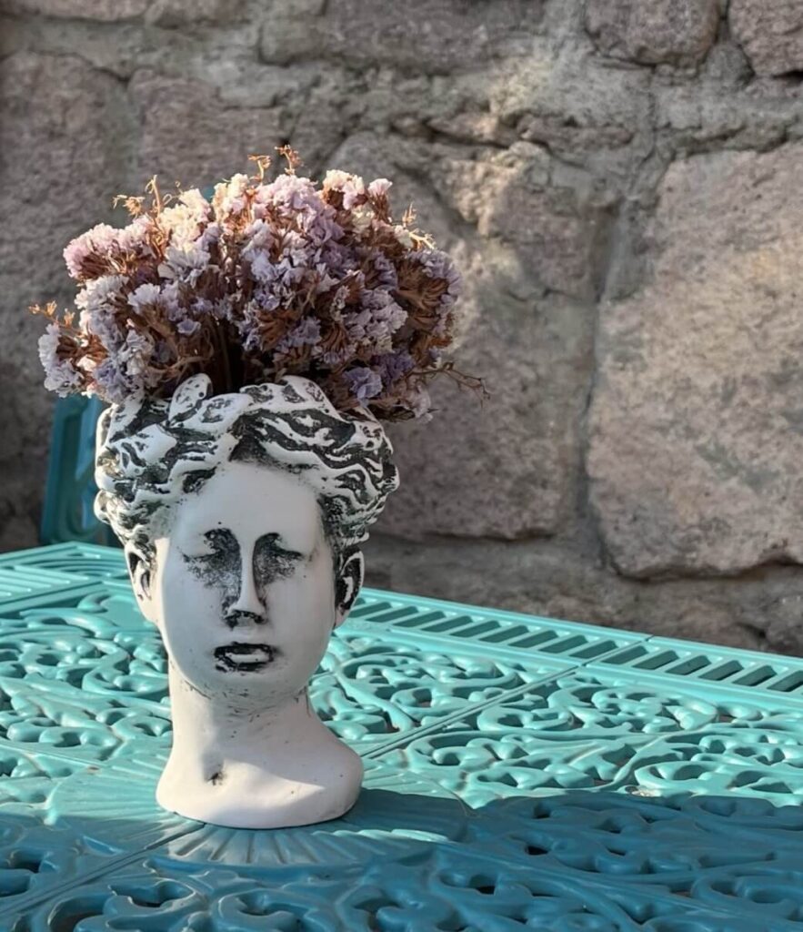 Frauenkopf Vase auf einem blauen Tisch. Im Hintergrund ist eine graue Steinwand zu sehen.